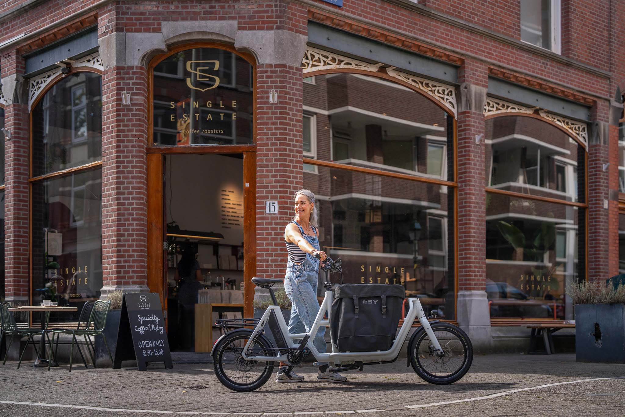Person überquert mit i:SY E-Bike die Straße vorm Single Estate Coffee in Den Haag – Szene aus einem Werbefilm für die Firma i:SY GmbH & Co. KG, produziert von Charlie Grashoff - The C/A Studio, Videoproduktion in Leipzig. Projekte deutschlandweit. Imagefilme, Corporate Videos, Brandfilme, Eventfilme, Industriefilme & mehr. Werbefilm Produktion. Fotografie Leipzig.
